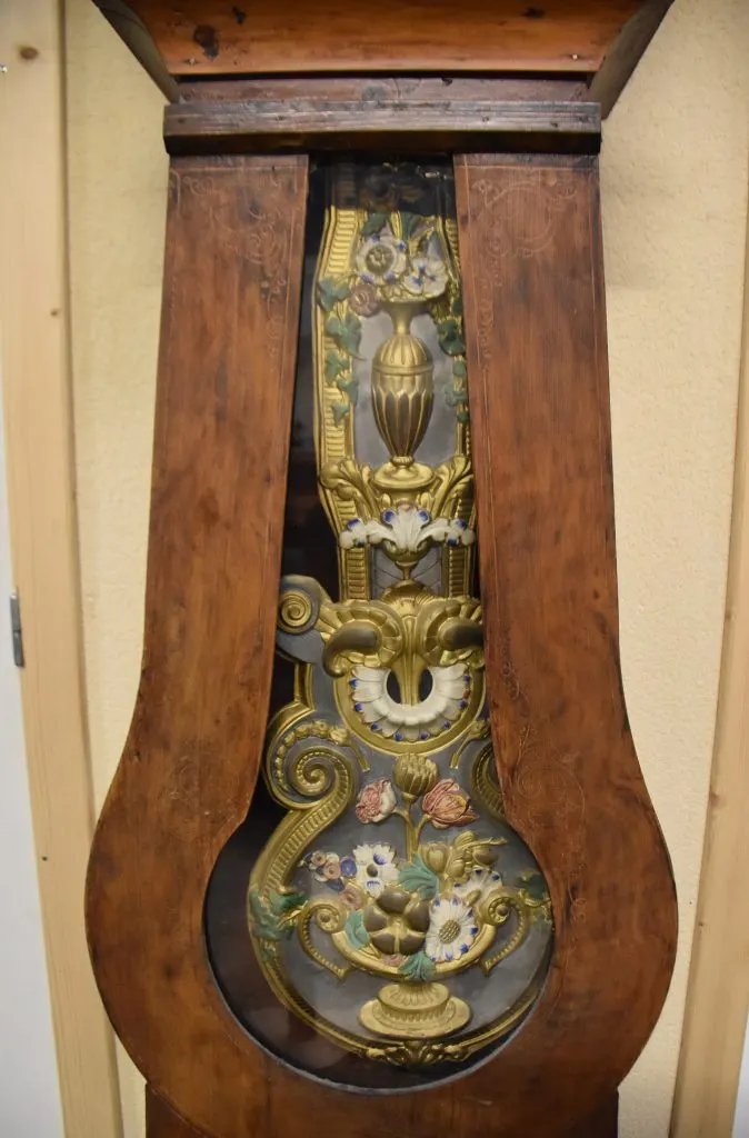 Balancier de comtoise en laiton repoussé et peint à décor de fleurs vu par la fenêtre d'une caisse en bois