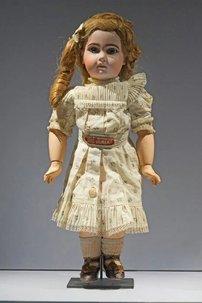Bébé Jumeau en pied, tête en biscuit, longs cheveux châtain clair, robe crème, présenté au Musée des Arts décoratifs