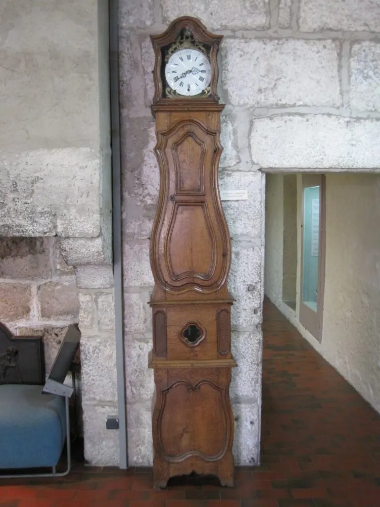 Horloge comtoise de Poligny à haute caisse en bois mouluré et galbé exposée contre un mur de pierre