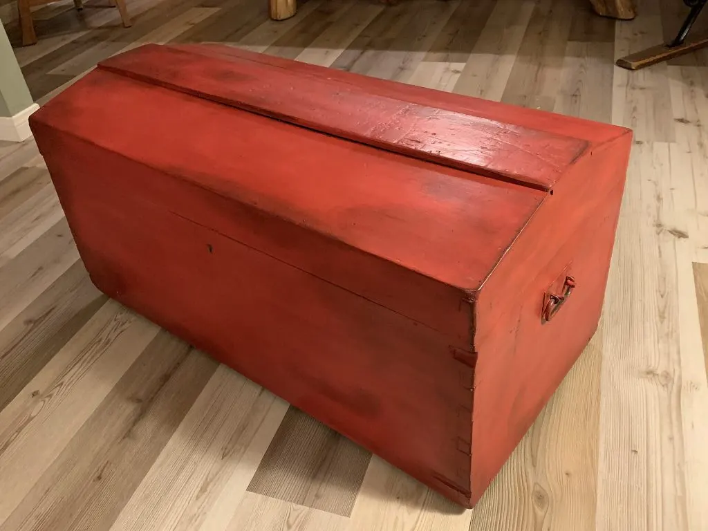 Malle de voyage repeinte en rouge à la peinture craie et cirée, posée sur un parquet clair