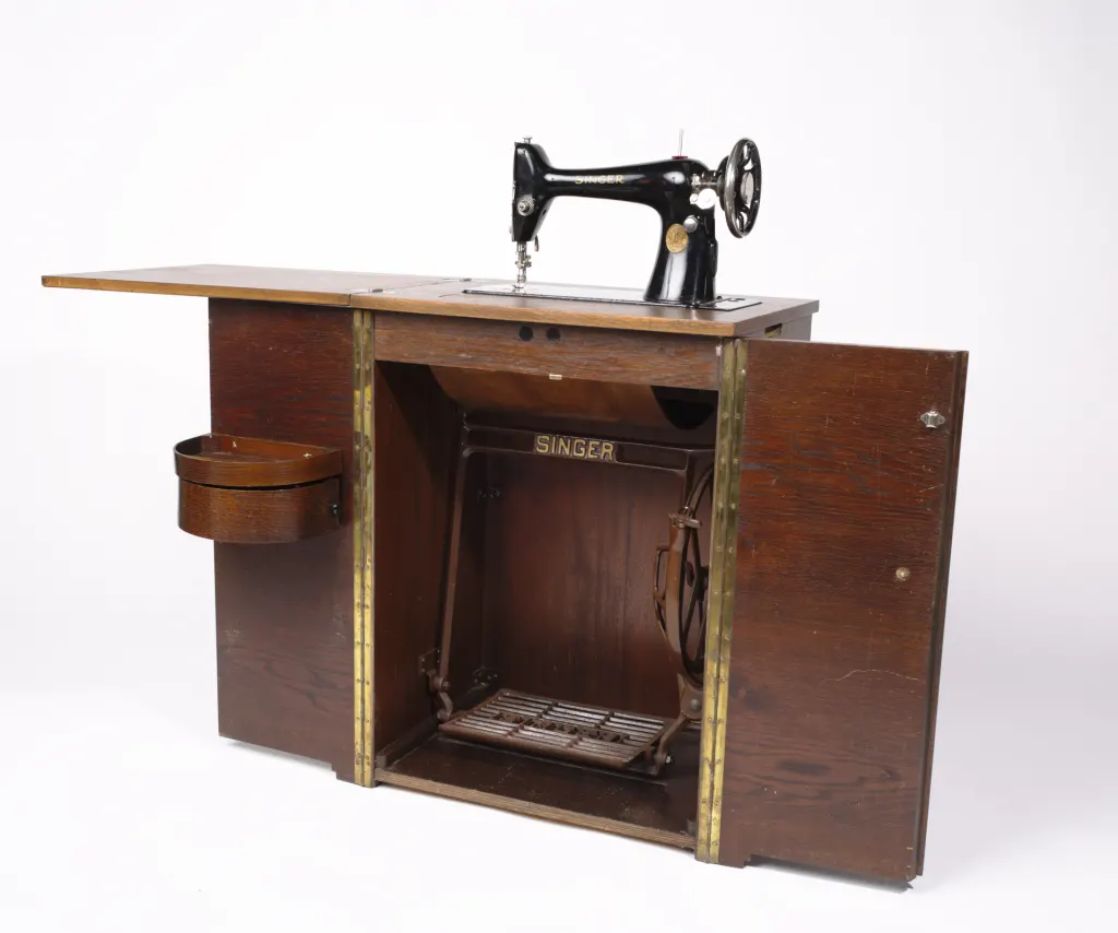 Meuble de machine à coudre Singer ouvert, tête noire sur le plateau, piétement en fonte et pédale visibles à l'intérieur