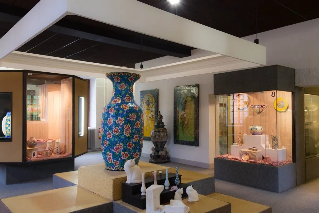 Salle du musée des émaux de Longwy avec un grand vase fleuri sur fond bleu au centre et des vitrines