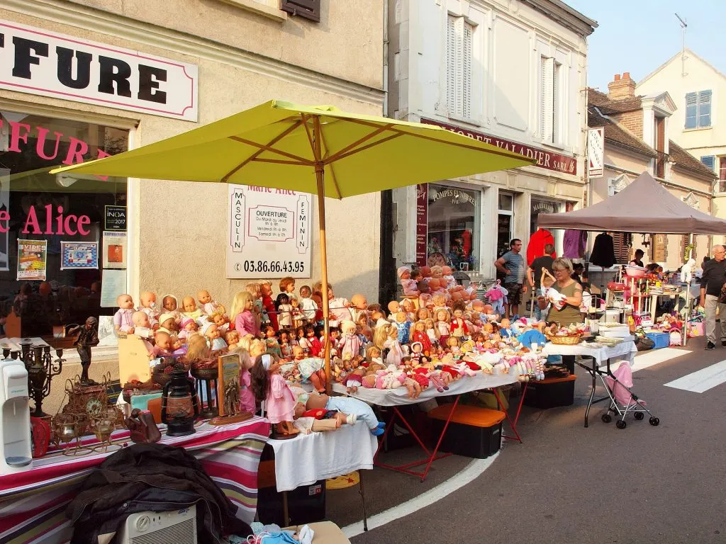 Stand de vide-greniers couvert de dizaines de poupées, sous un parasol vert, dans une rue de Villeneuve-la-Guyard